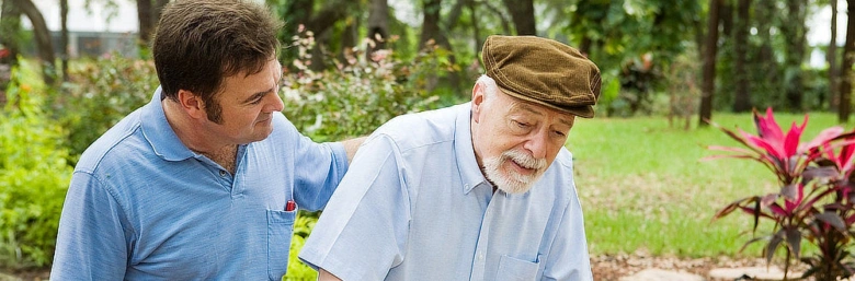 Mladý muž pomáhá staršímu muži s parkinsonovou chorobou v zahradě.