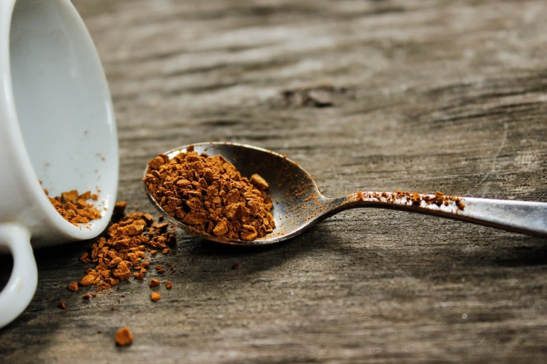 Lžíce s granulovanou instantní kávou vedle převrácené šálku na dřevěném povrchu.