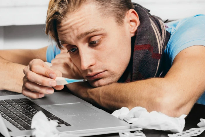 Muž s teploměrem u notebooku vypadá nemocně.