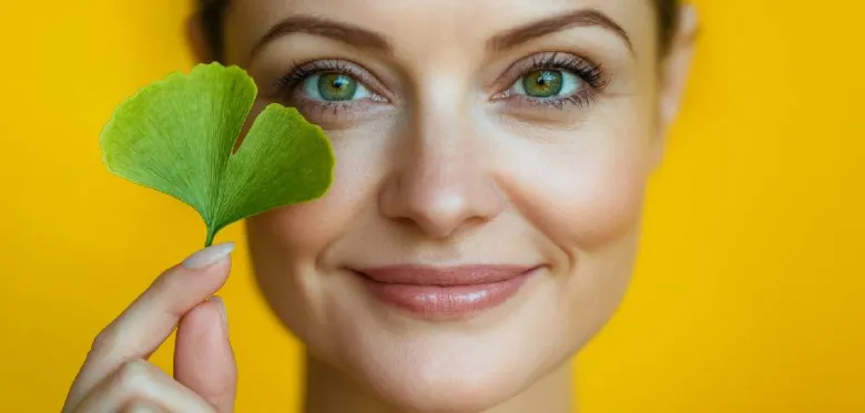 Žena drží list ginkgo biloba u oka na žlutém pozadí.