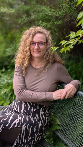 Žena s kudrnatými vlasy sedí venku v zeleném prostředí.