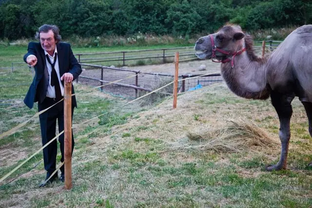 Bolek Polívka stojí vedle velblouda na trávnate ploše.