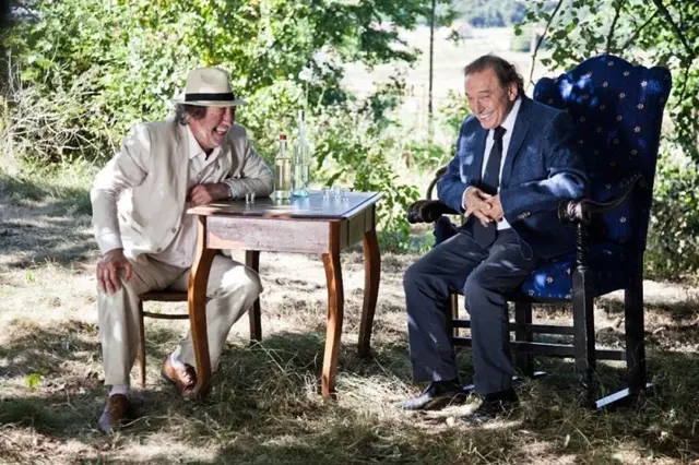 Bolek Polívka sedí venku s přítelem u stolu.