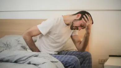 Muž sedí na posteli a drží si hlavu v bolesti.