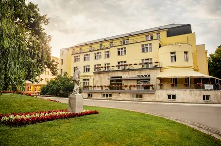 Fotografie žlutého lázeňského domu s sochou na trávníku.