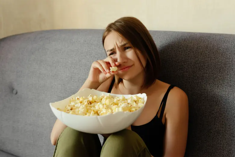 Žena sedí na gauči s miskou popcornu a vypadá smutně.