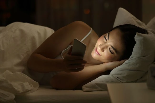 Žena leží v posteli s mobilním telefonem ve tmě.