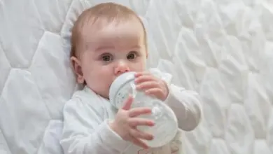Babička pije z lahvičky ležící na bílém povlečení.