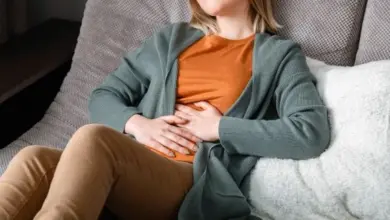 Žena sedí na gauči a drží si břicho kvůli bolesti.